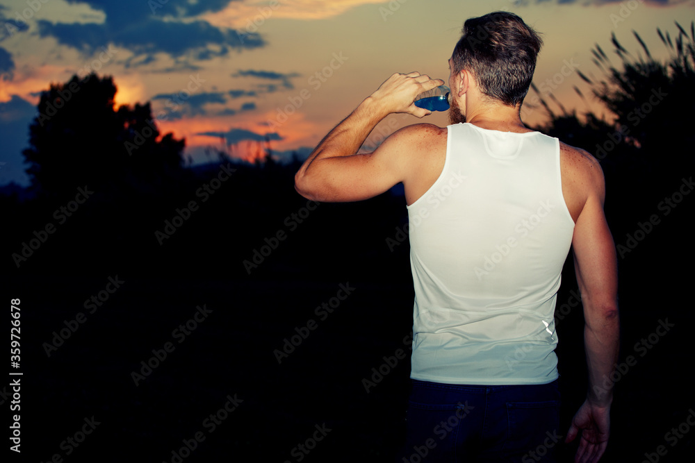 Young athlete with muscular body drinking water from a bottle while enjoying the sunset with copy space area for your text message or content, male runner resting after intensive evening jog outdoors
