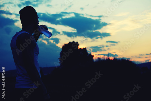 Silhouette of male jogger drinking water while enjoying the sunset with copy space area for your text message or content, muscular build sportsman taking break after physical exercises in the park