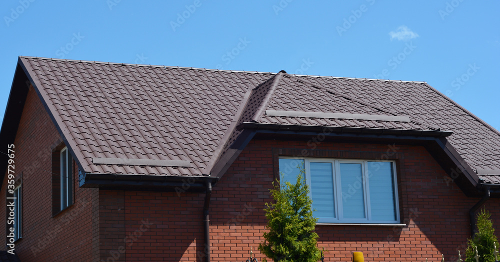 A close-up on a brown attic mansard metal tiled roof with snow guards, snow stoppers of a brick house against blue sky. A close-up on a brown attic mansard metal tiled roof with snow guards, snow stoppers of a brick house against blue sky.