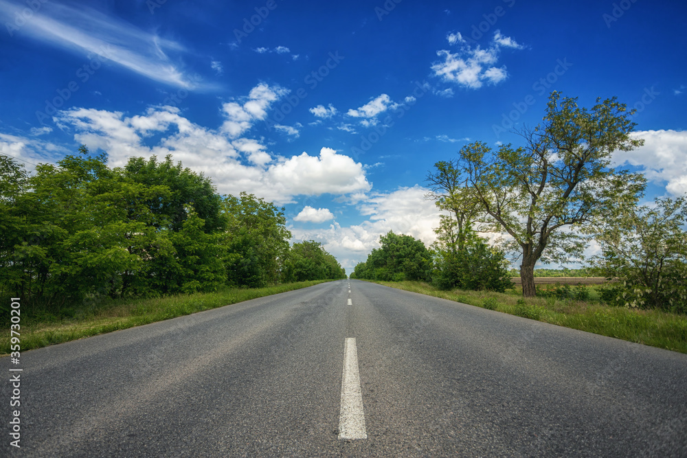 Car-free, empty asphalt country road, highway, on a sunny summer, spring day, receding into the distance, against a blue sky with white clouds and trees on the side of the road