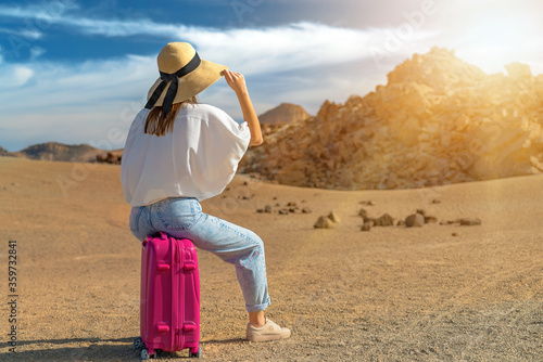 Girl in a white shirt and hat sit on a pink suitcase on a volcanic sandy scenery. Teide volcano, Tenerife, Spain.