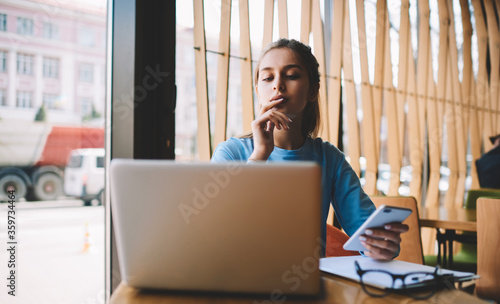 Concentrated talented student dressed in casual outfit carefully reading information on websites via macbook computer.Young hipster girl holding smartphone while looking at modern laptop in cafe
