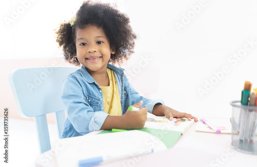 Portrait of smiling african girl sitting at desk in living room and looking at camera while drawing at home.New normal.Covid-19 coronavirus.Social distancing.stay home