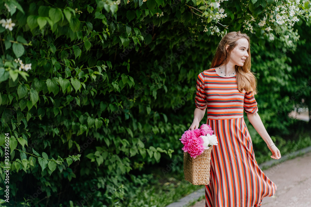 Outdoor portrait of thoughtful romantic woman wearing pink strip dress, holding straw wicker bag with peony flowers and hat, walking in green park outdoor. Copy, empty space for text. Outdoor portrait of thoughtful romantic woman wearing pink strip dress, holding straw wicker bag with peony flowers and hat, walking in green park outdoor. Copy, empty space for text.