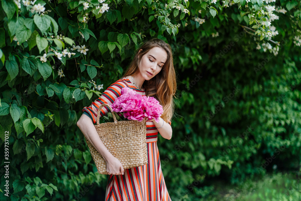Outdoor portrait of young happy smiling young girl wearing pink strip dress, holding straw wicker bag with peony flowers and hat, posing in green park outdoor. Copy, empty space for text. Outdoor portrait of young happy smiling young girl wearing pink strip dress, holding straw wicker bag with peony flowers and hat, posing in green park outdoor. Copy, empty space for text.