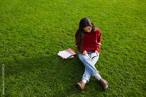 Pretty student girl sitting on the grass reading a book outdoors, young attractive girl with brunette long hair seating on the copy space grass background holding some book in the hands