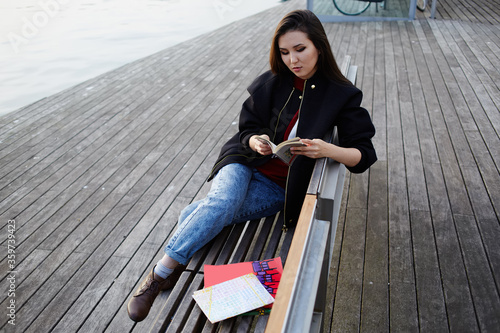 Attractive female student reading a book outdoors, beautiful girl holding an open book sitting on wooden bench near artificial lake, tourist woman resting after walk in new city reading some book photo