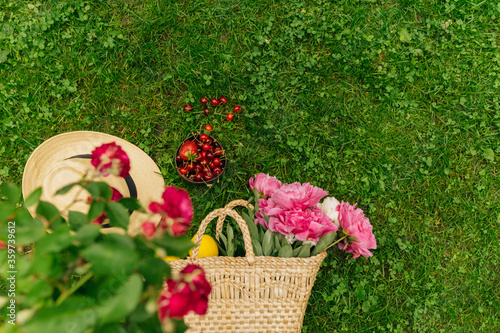 Wicker bag with peony flowers, straw hat, bowl of strawberries and cherries, glass bottle, lemons on green lawn grass, outdoor, Summer vacation, simple set for picnic. Spring vacation mood concept.