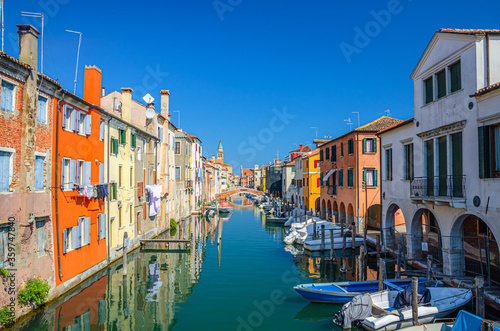 Fototapeta Naklejka Na Ścianę i Meble -  Chioggia cityscape with narrow water canal Vena with moored multicolored boats between old colorful buildings and brick bridge, blue sky background in summer day, Veneto Region, Northern Italy