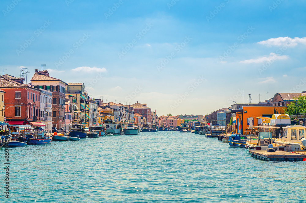 Obraz premium Narrow water canal with moored boats and fishing industrial ships and old buildings on embankment in Chioggia town historical centre, blue sky background in summer day, Veneto Region, Northern Italy