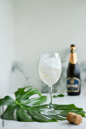 Sparkling wine with ice in glass standing on the green mostera leaf on the white table and champagne bottle