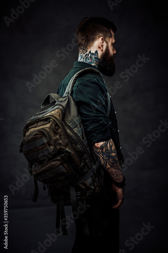 Travel and education concept. Back view of a bearded tattooed hipster guy with backpack in a dark studio