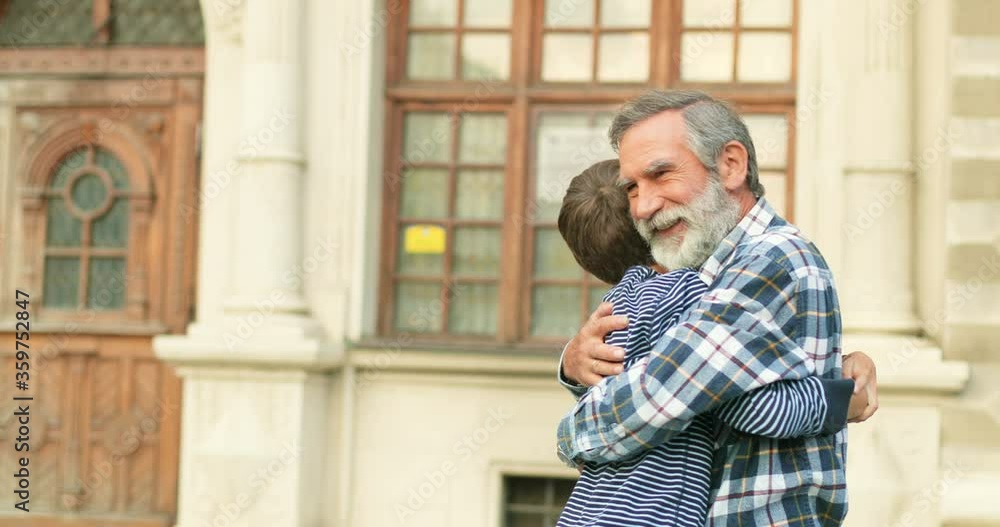 Caucasian happy senior grandfather meeting and hugging his cute joyful teen grandson at street in city. Cheerful old gray-haired man and teenage boy meet outdoor. Caucasian happy senior grandfather meeting and hugging his cute joyful teen grandson at street in city. Cheerful old gray-haired man and teenage boy meet outdoor.