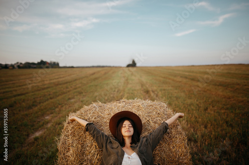 Valokuva Stylish girl relaxing on hay bale in summer field in sunset
