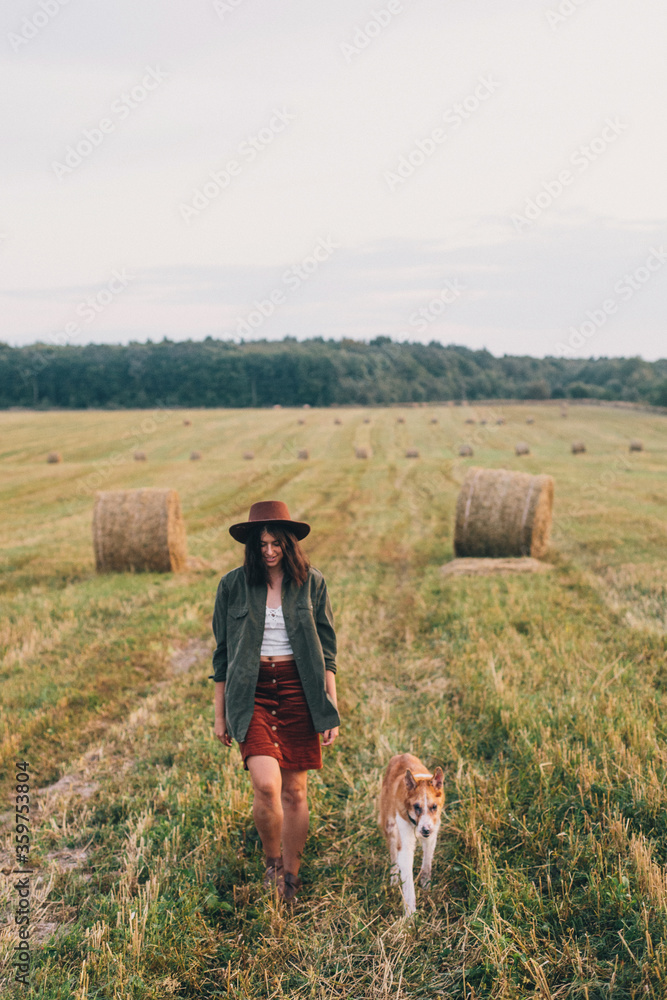 Stylish girl walking with her dog at hay bale in summer field in sunset. Young woman in hat traveling with pet in haystack field. Countryside slow life
