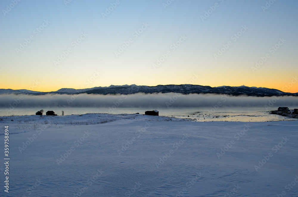 dense ice fog in winter over fjord and mountain landscape at colorful sunset
