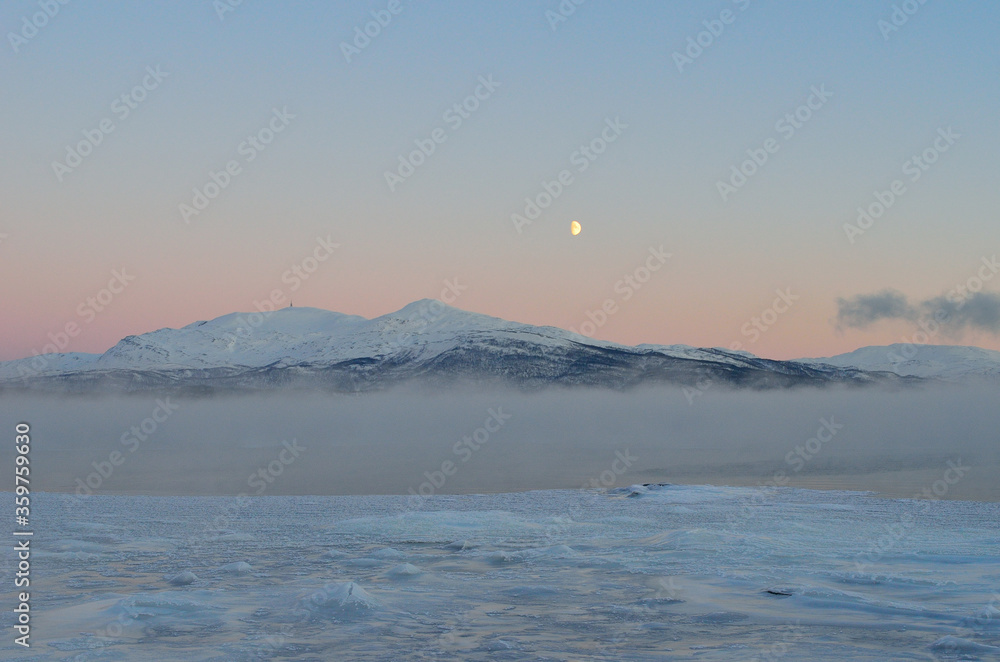 pink sunset sky and full moon over winter fjord with dense ice fog and reflection on water surface