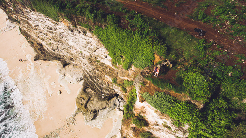 Top view aerial photo from flying drone of a young loving couple are enjoying amazing landscape of Indian Ocean  while are standing on a green cliff during their beautiful honeymoon in Bali Indonesia