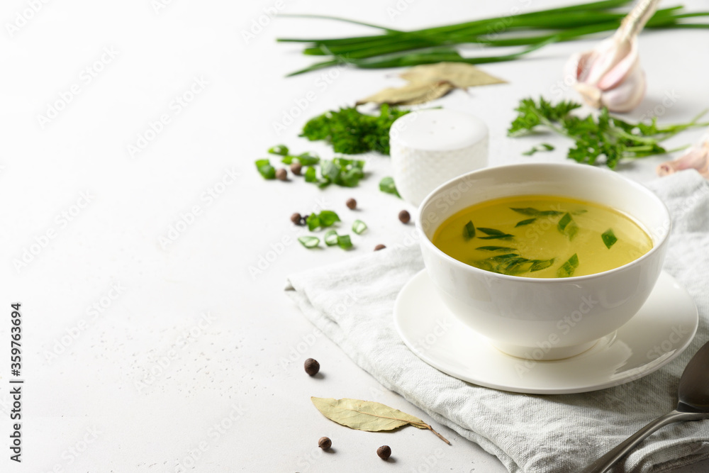 Chicken broth with spices, greens, garlec, parsley, onion in white bowl on white table. Copy space. Close up. Chicken broth with spices, greens, garlec, parsley, onion in white bowl on white table. Copy space. Close up.