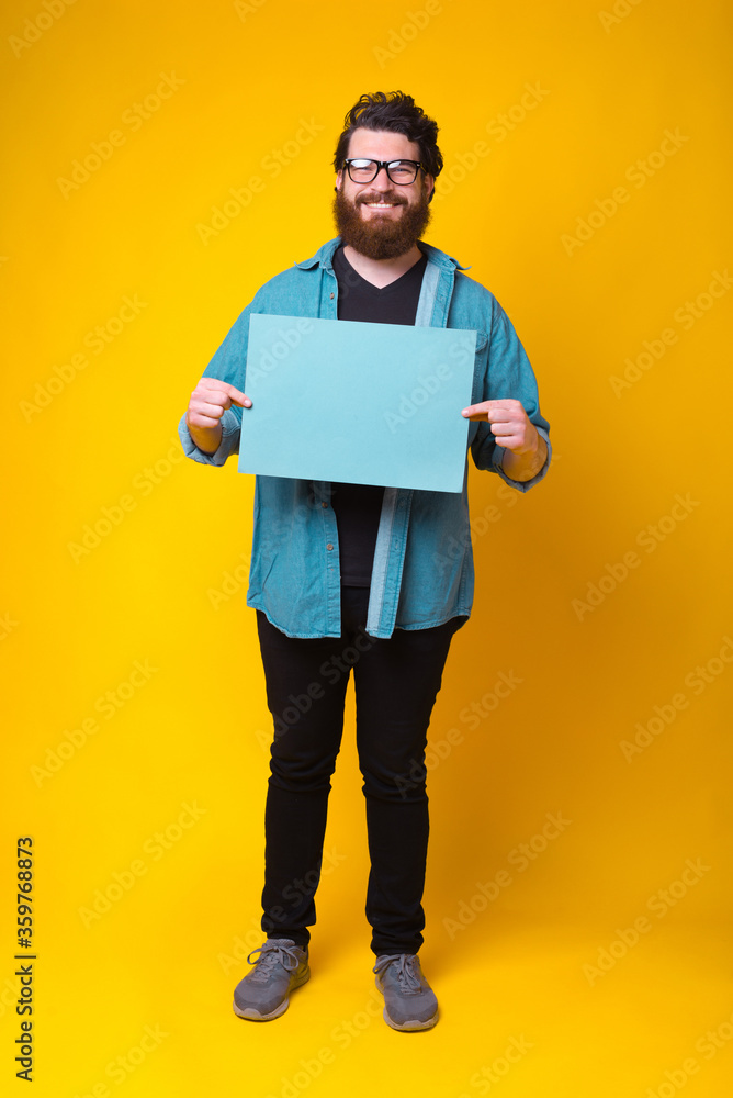 Smiling at the camera hipster is holding a blue paper while standing over yellow background Smiling at the camera hipster is holding a blue paper while standing over yellow background