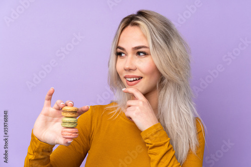 Teenager girl over isolated purple background holding colorful French macarons and thinking an idea