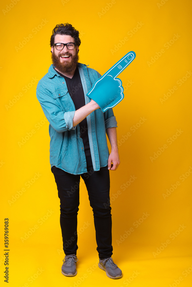 Handsome bearded man is wearing a fan foam glove standing on yellow background. Handsome bearded man is wearing a fan foam glove standing on yellow background.