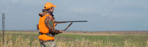 Fotografia A man with a gun in his hands and an orange vest on a pheasant hunt in a wooded area in cloudy weather