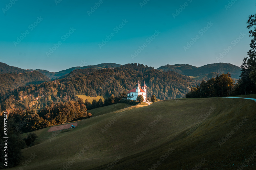 Catholic church on hilltop illuminated by sun. Meadow grassland with tractor on farming field in shadow. In background hills covered with forest trees. Late summer evening in Slovenia Catholic church on hilltop illuminated by sun. Meadow grassland with tractor on farming field in shadow. In background hills covered with forest trees. Late summer evening in Slovenia