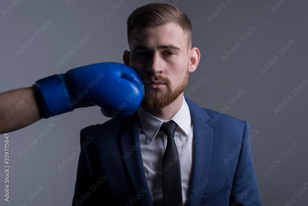 Naklejka premium close-up portrait of a young bearded guy in a business suit, businessman, side view the hand of an opponent in a boxing glove strikes. The moment of punching in the face.