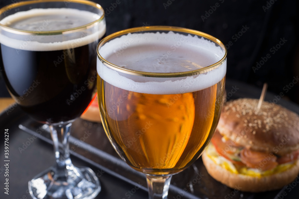 close-up, beer glasses with craft dark and light beer with foam, against a background of burgers, with a variety of ingredients, including shyomga, beef meat patty, cheese burger. In a rustic style close-up, beer glasses with craft dark and light beer with foam, against a background of burgers, with a variety of ingredients, including shyomga, beef meat patty, cheese burger. In a rustic style