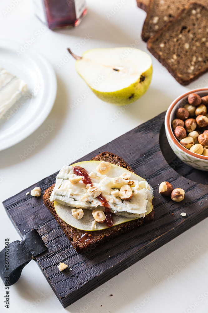 Open sandwich with rye bread, fresh pear , gorgonzola cheese, hazelnuts and cranberry sauce and bowl with nuts on wooden board and sandwich ingredients on white background. Open sandwich with rye bread, fresh pear , gorgonzola cheese, hazelnuts and cranberry sauce and bowl with nuts on wooden board and sandwich ingredients on white background.