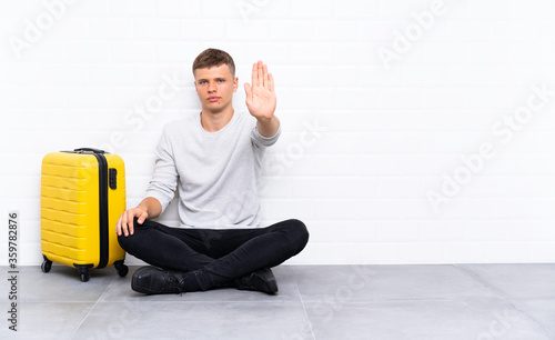 Young handsome man sitting on the floor with a suitcase making stop gesture with her hand