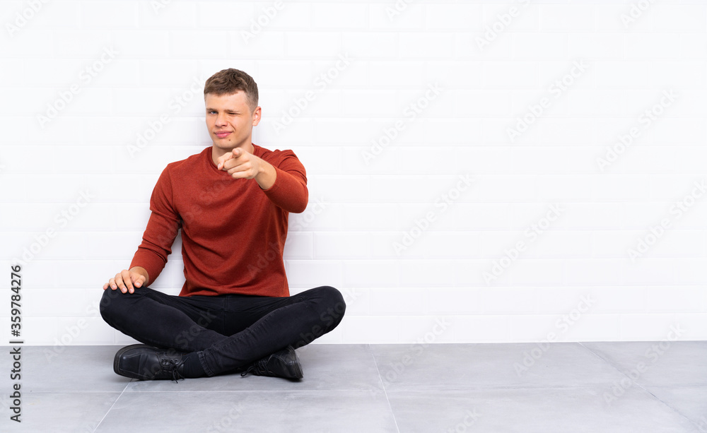 Young handsome man sitting on the floor points finger at you