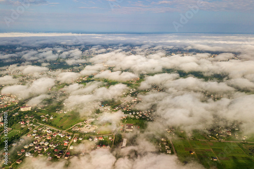 Aerial view of white clouds above a town or village with rows of buildings and curvy streets between green fields in summer. Countryside landscape from above.