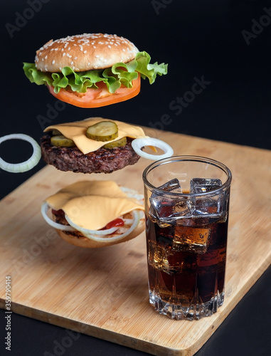 Delicious Burger with floating ingredients on the background of a wooden table. Bun with sesame seeds. Juicy meat cutlet. Carbonated brown drink with ice.