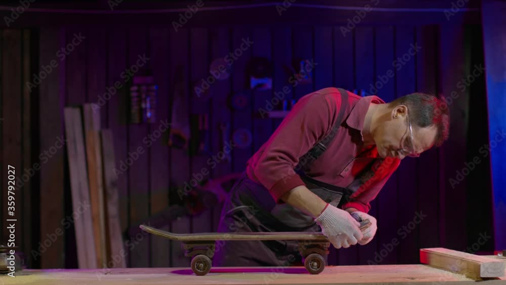 Caucasian adult male carpenter in overalls and goggles repairs a skateboard in his workshop. Making craft skateboards concept. Men's work. Stylish workshop lighting. Garage for work. Caucasian adult male carpenter in overalls and goggles repairs a skateboard in his workshop. Making craft skateboards concept. Men's work. Stylish workshop lighting. Garage for work.