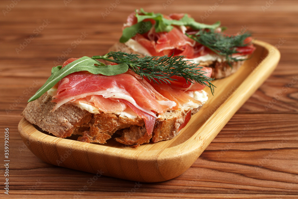 Buckwheat toasts with goat cheese, ham and arugula lying on a bamboo plate on a wooden table. Buckwheat toasts with goat cheese, ham and arugula lying on a bamboo plate on a wooden table.