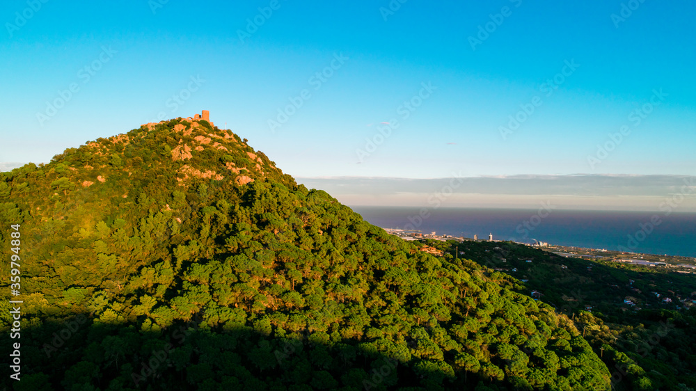 Landscape with a mountain full of green trees on the left with a building on the top and on the right to a lower level and far away the dark blue of the sea with the clear blue sky.