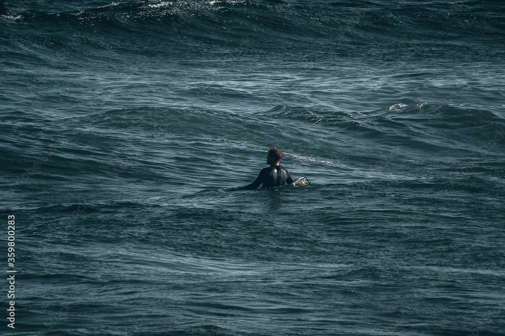 Silhouette of surfer from behind sitting on his surfboard out in the ocean waiting for a wave to catch Silhouette of surfer from behind sitting on his surfboard out in the ocean waiting for a wave to catch