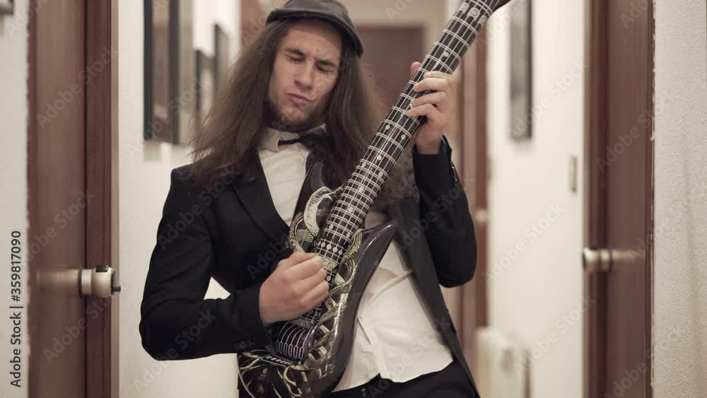 Young long-haired man in suit plays inflatable toy guitar in apartment Young long-haired man in suit plays inflatable toy guitar in apartment
