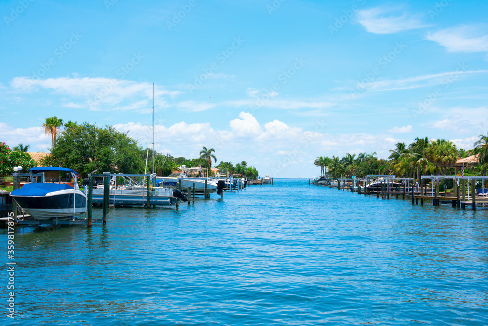 Residential saltwater canal in south Florida with beautiful blue water and a variety of boats at docks and on electric lifts with green palm trees lining the shoreline on a bright sunny afternoon.