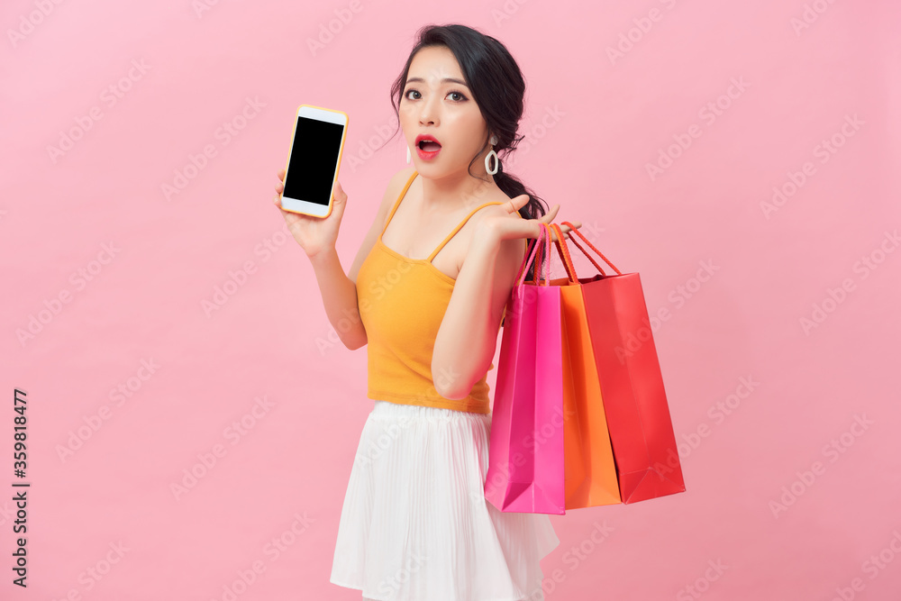 Photo of shocked woman 20s in dress looking at smartphone in hand with surprise while holding shopping bags isolated over pink background Photo of shocked woman 20s in dress looking at smartphone in hand with surprise while holding shopping bags isolated over pink background