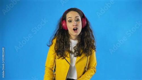 Young woman in yellow jacket dancing while listening music on headphones in Studio with blue Background.