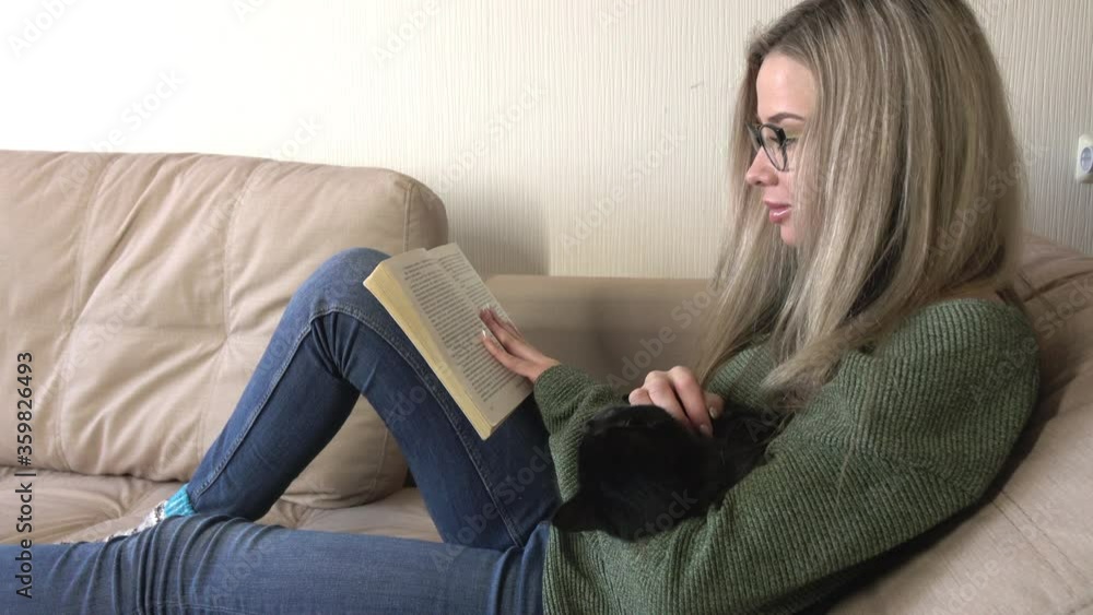 A pretty young woman is happy to read a book at home. The black kitten watches curiously A pretty young woman is happy to read a book at home. The black kitten watches curiously