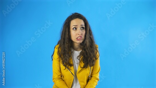 Nervous beautiful Caucasian female with long brown hair rubbing hands gnaw nails in Studio with blue Background.