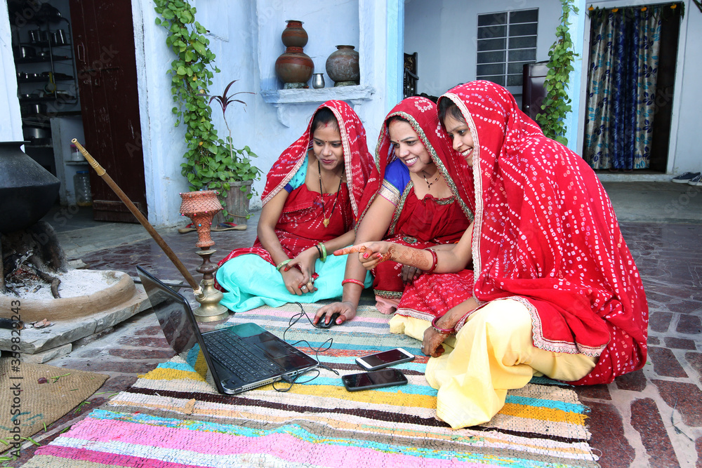 Traditional Indian young married women working in traditional kitchen on laptop. Using technology in rural households. Women cook food and learn technology. Rural women using Laptop.