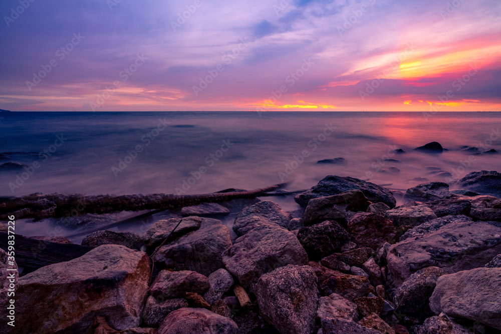 Stone beach and calm sea at dusk with purple sunset sky. Tropical sea. Skyline in the evening with golden, purple, and red sky. Seascape. Summer vacation travel. Rock coast. Beauty in ocean nature.
