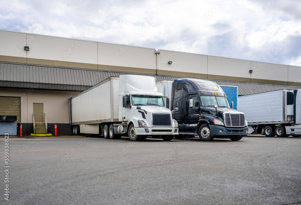 Fototapeta premium Black and white big rigs semi trucks with semi trailers loading cargo at warehouse dock with gates for each