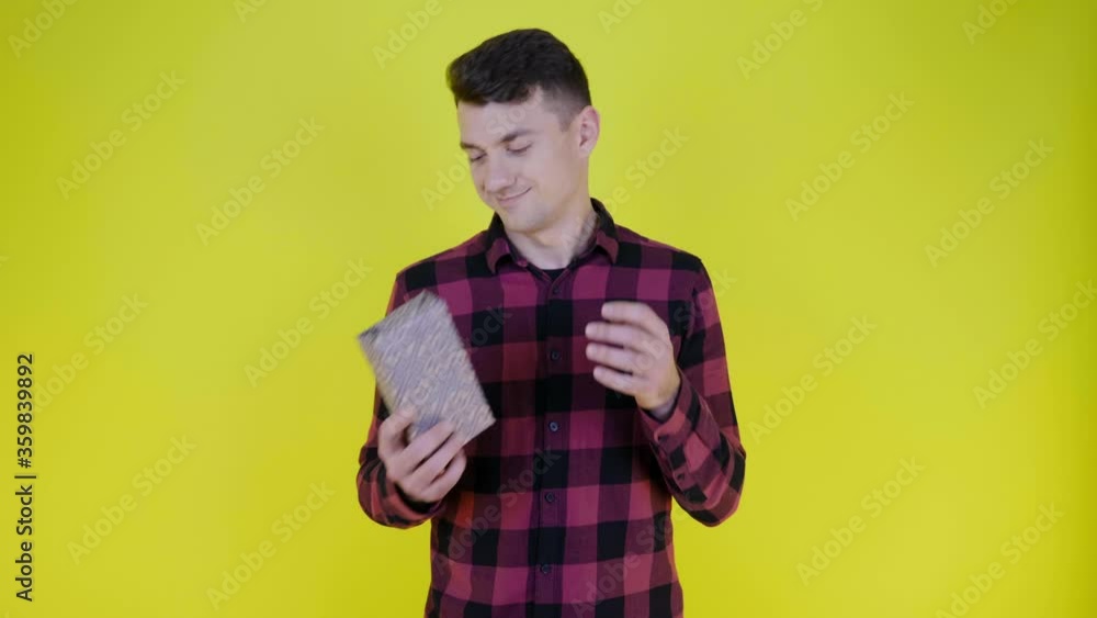 Handsome man in a pink plaid shirt throws box with gift from hand to hand on yellow background with copy space. Place for text or product. 4K slow motion footage Handsome man in a pink plaid shirt throws box with gift from hand to hand on yellow background with copy space. Place for text or product. 4K slow motion footage