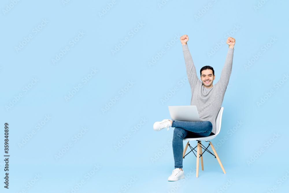 Portrait of happy young handsome Caucasian man sitting using laptop doing arms raised winning gesture in isolated studio blue background Portrait of happy young handsome Caucasian man sitting using laptop doing arms raised winning gesture in isolated studio blue background